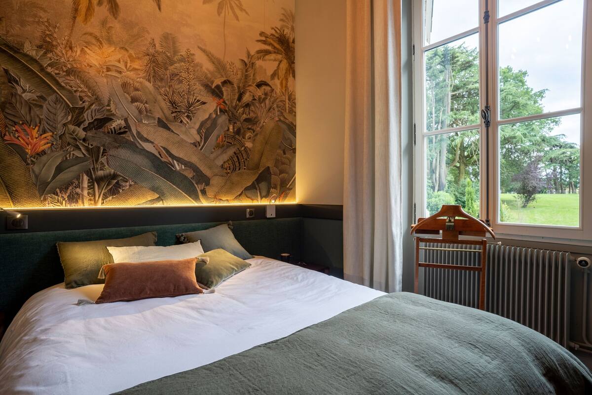 A bed with a green headboard, a mural on the wall, and a window with a view of greenery.