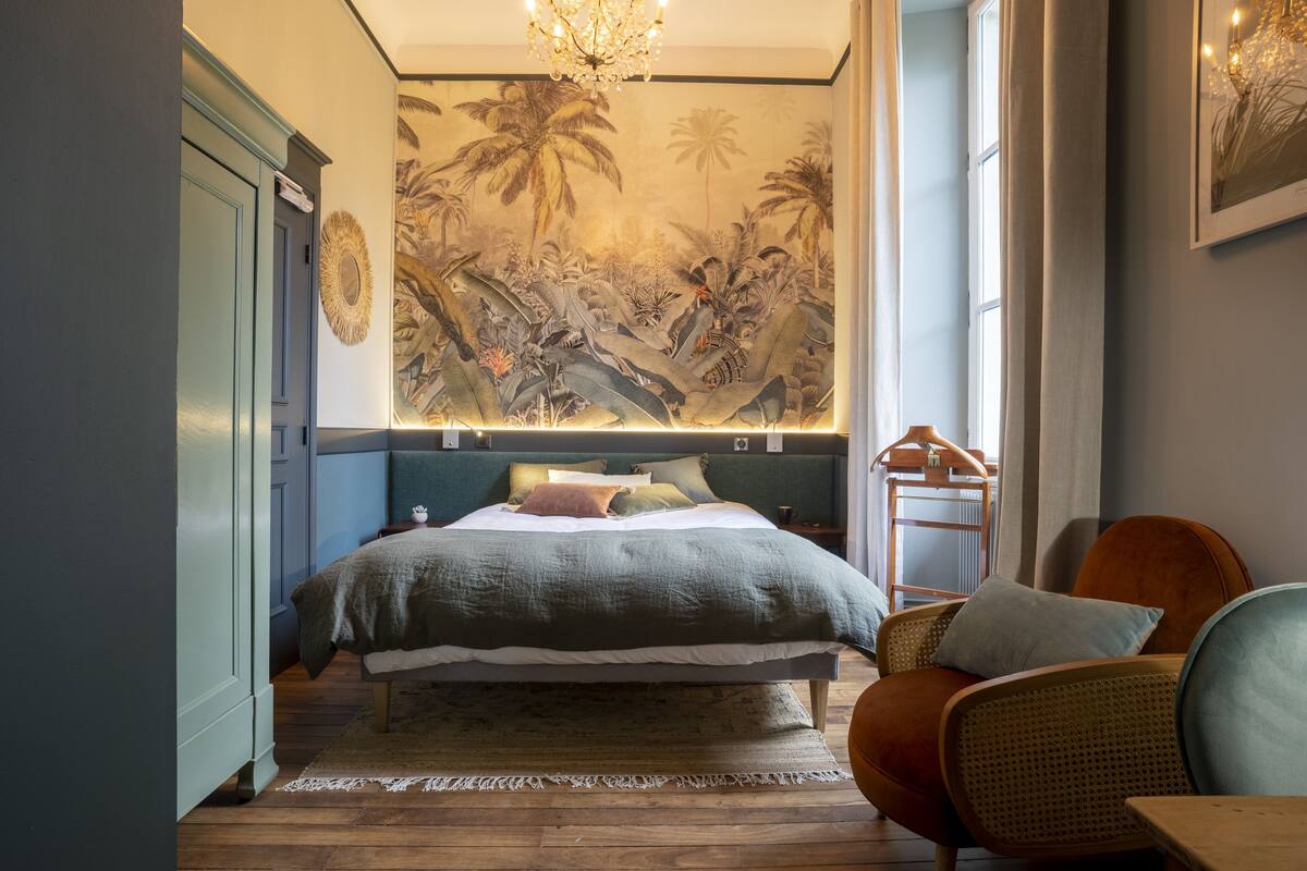 A bedroom with a large bed, a rattan chair, a wooden nightstand, and a wall mural.