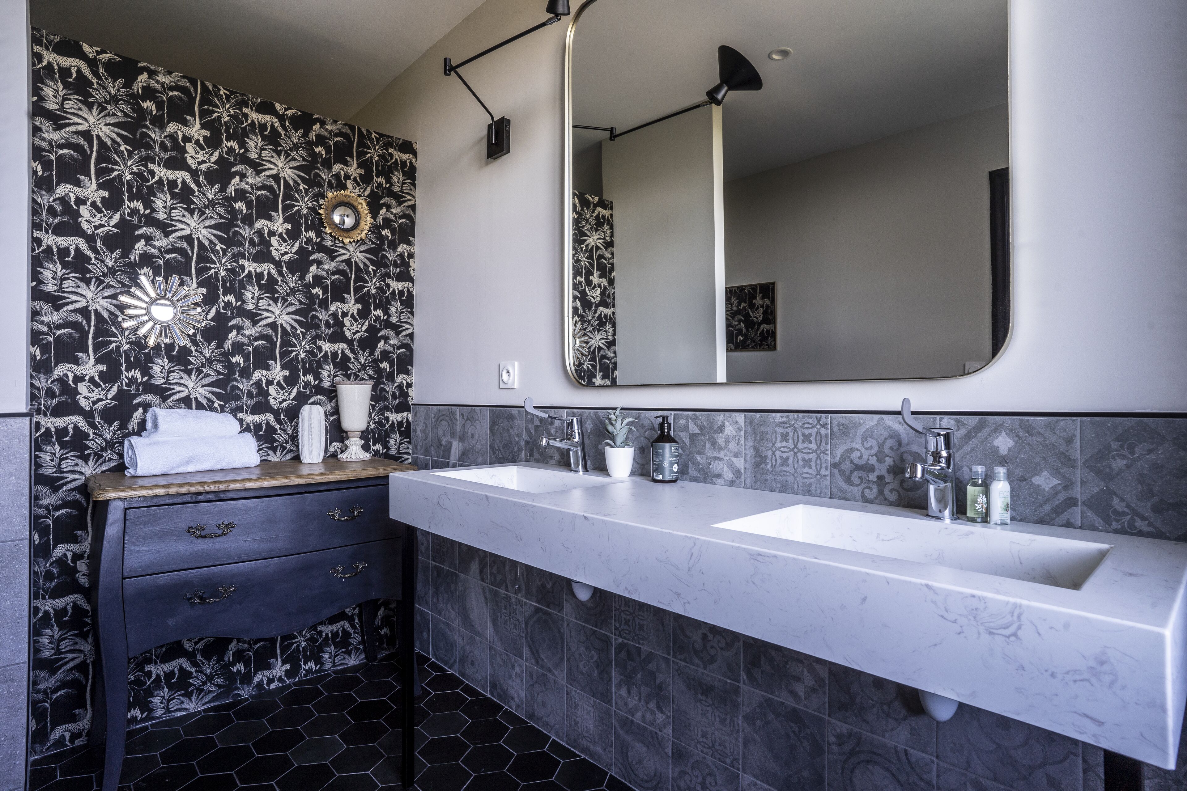 Een badkamer met behang in zwart-wit bloemenmotief, twee wasbakken, een spiegel en een wastafelmeubel met lades.