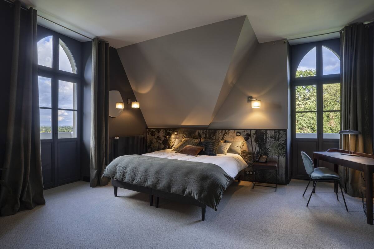 A bedroom with a large bed, two bedside tables, a desk, and a chair. There are two windows with curtains, and the room has a sloped ceiling.