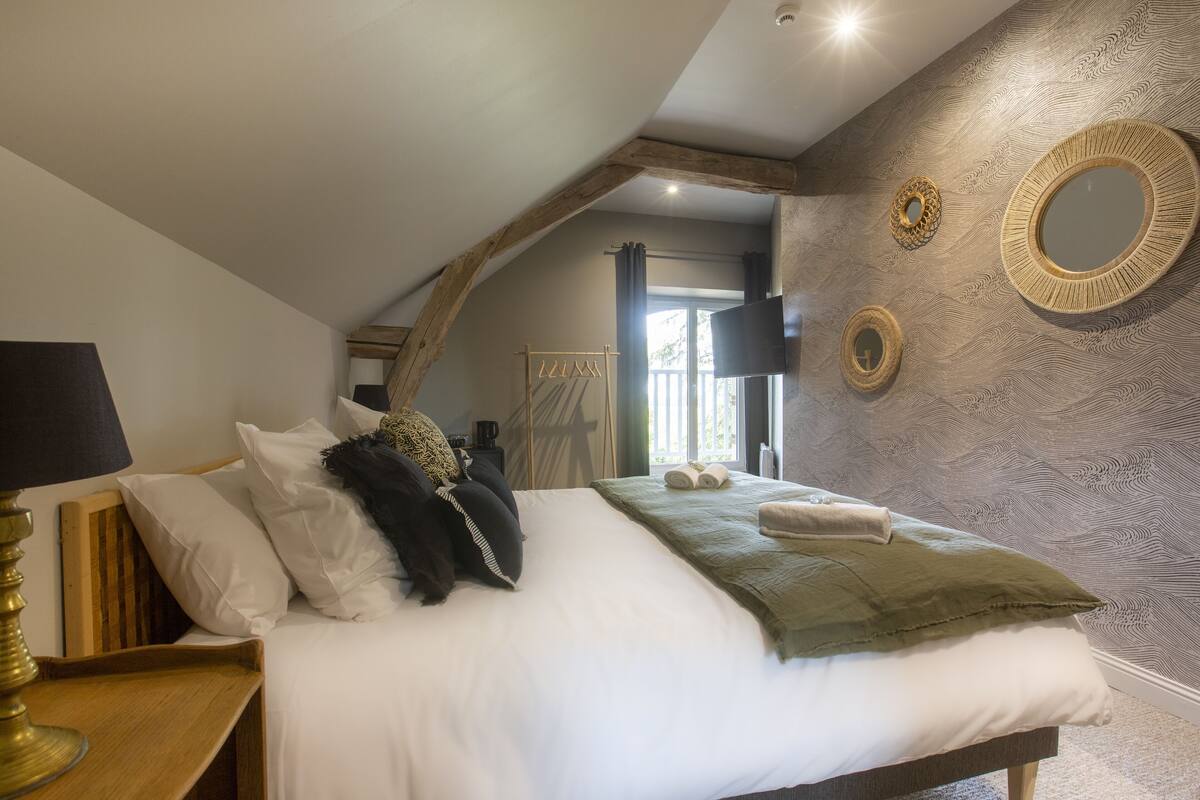 A bedroom with a bed, a bedside table with a lamp, a mirror, and a window with curtains.