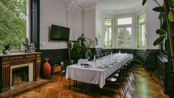 A long dining table set for a meal in a room with a fireplace, large windows, and a television on the wall.
