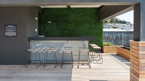 A rooftop bar with a green wall, bar stools, and a view of buildings in the background. - Brand New Modern De Waterkant Apartment (Cape Town)