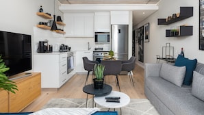 A modern living room with a grey sofa, a coffee table, and a TV. There is a kitchen area with white cabinets, a microwave, and a refrigerator. - Brand New Modern De Waterkant Apartment (Cape Town)