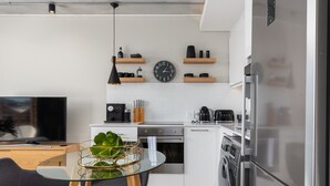 A modern kitchen with white cabinets, a stainless steel refrigerator, a built-in oven, and a coffee maker. There is a round table with chairs and a potted plant. - Brand New Modern De Waterkant Apartment (Cape Town)