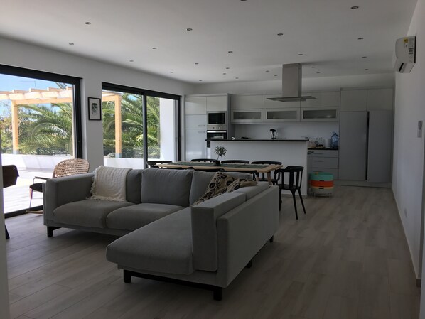 Una cocina moderna con armarios blancos, una zona de comedor con una mesa y sillas, y un sofá seccional gris en un amplio salón con ventanas grandes.