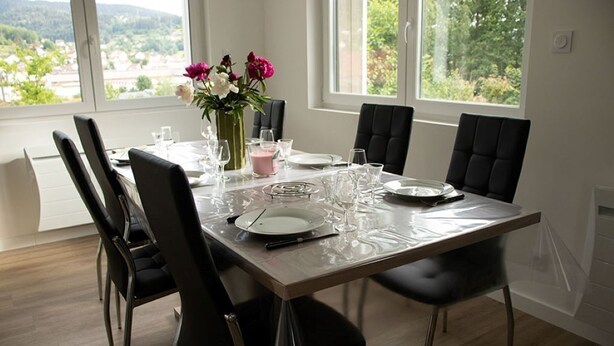 A dining table set for six with black chairs, a vase with flowers, and a view of the outdoors through large windows. - Quiet apartment with terrace for 6 people, 3 bedrooms. (Gérardmer)