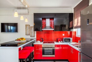 Eine moderne Küche mit roten Schränken, einem Edelstahlkühlschrank, einer Mikrowelle und einem eingebauten Backofen. Über der Kochinsel hängen drei Pendelleuchten, auf ihr befindet sich eine Obstschale und eine Kaffeemaschine.