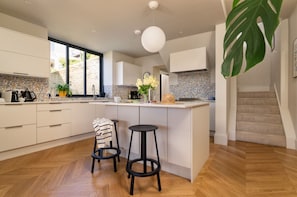 A modern kitchen with white cabinets, a large island with stools, a hanging light fixture, and a view of a stone wall outside.