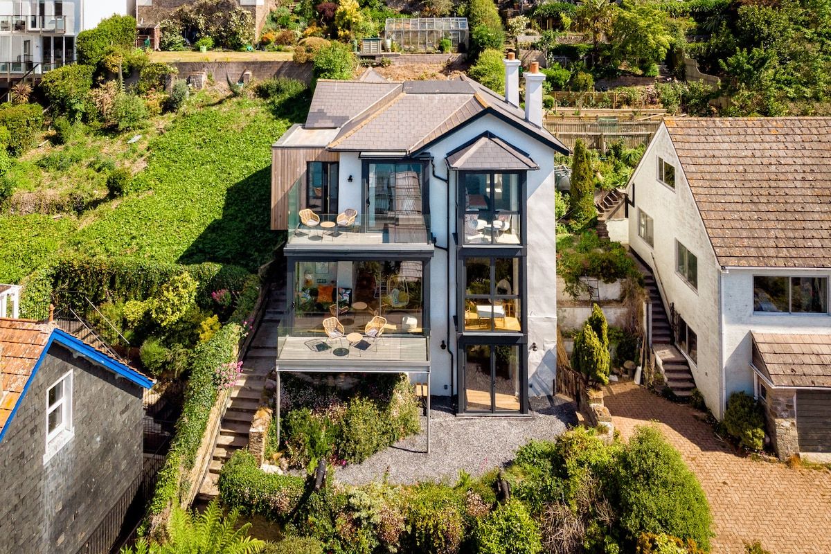 A modern two-story house with a glass balcony, surrounded by lush greenery and a steep hillside.