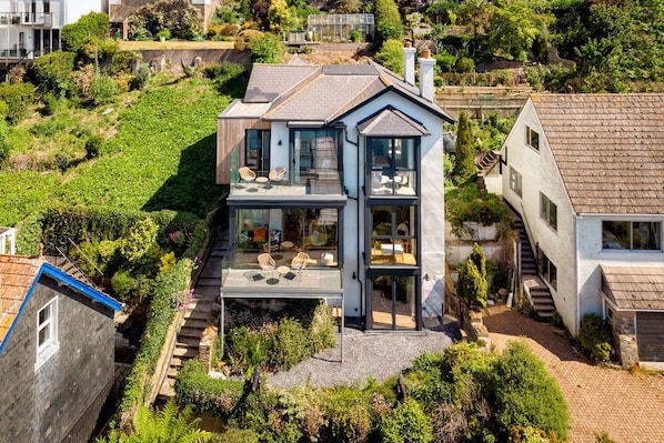 A modern two-story house with a glass balcony, surrounded by lush greenery and a steep hillside.