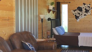 Interno di una baita in legno con un divano in pelle, un letto, un termosifone e una pelle di mucca appesa al muro.