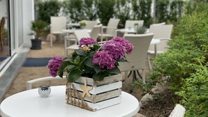 A white outdoor seating area with wicker chairs and a round table, a wooden planter box with a star decoration, and a potted plant with purple flowers. - Pension am Meer (Ostseebad Baabe)
