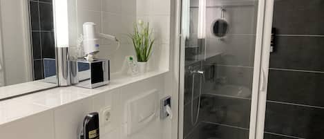 Um banheiro moderno com secador de mãos, um pequeno vaso de plantas, chuveiro com proteção de vidro e pia branca.