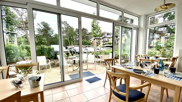 A dining area with large windows, wooden tables, and chairs. - Pension am Meer (Ostseebad Baabe)