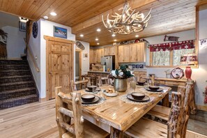 A rustic interior with a wooden dining table, chairs, a chandelier, and a kitchen area.