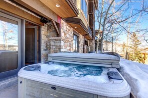 A hot tub outside a stone building with a wooden deck and a glass door.