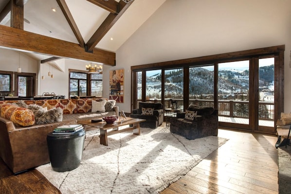 Un vaste salon doté d’une grande fenêtre offrant une vue sur les montagnes, de poutres en bois au plafond et d’un coin salon confortable agrémenté de coussins à motifs.