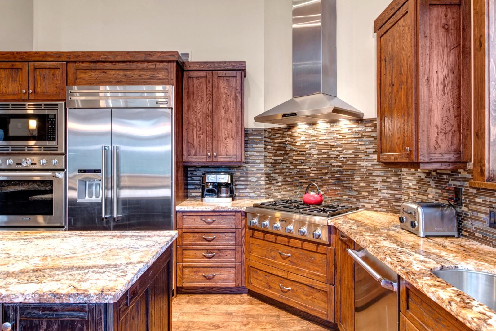 A kitchen with stainless steel appliances, wooden cabinets, a granite countertop, and a backsplash with a mosaic tile pattern.