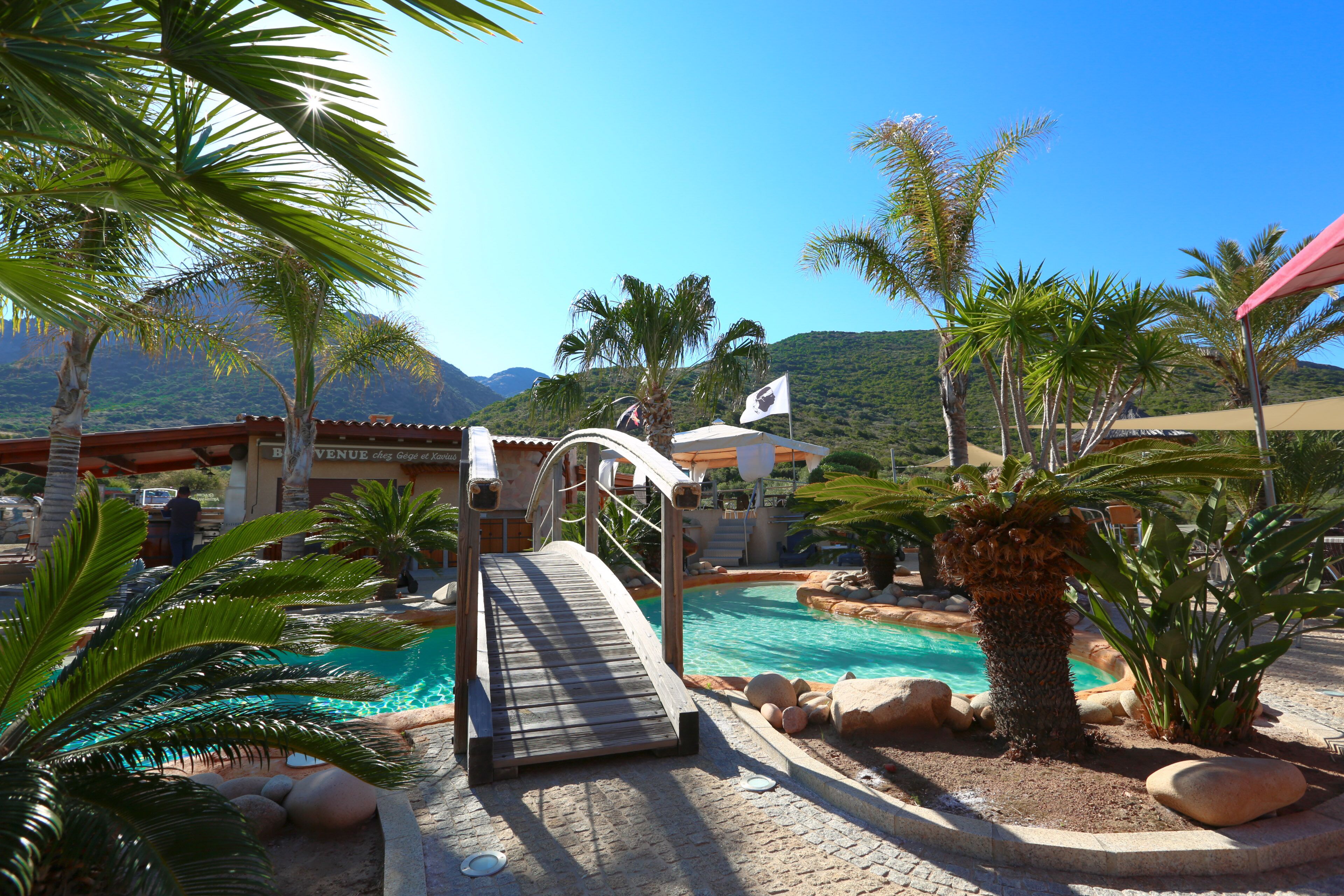 Zona de piscina con un puente de madera, palmeras y montañas al fondo.