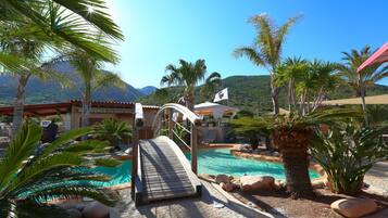 Zona de piscina con un puente de madera, palmeras y montañas al fondo.