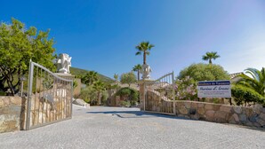 Una entrada con rejas, muros de piedra, un cartel que indica residencia vacacional y palmeras.