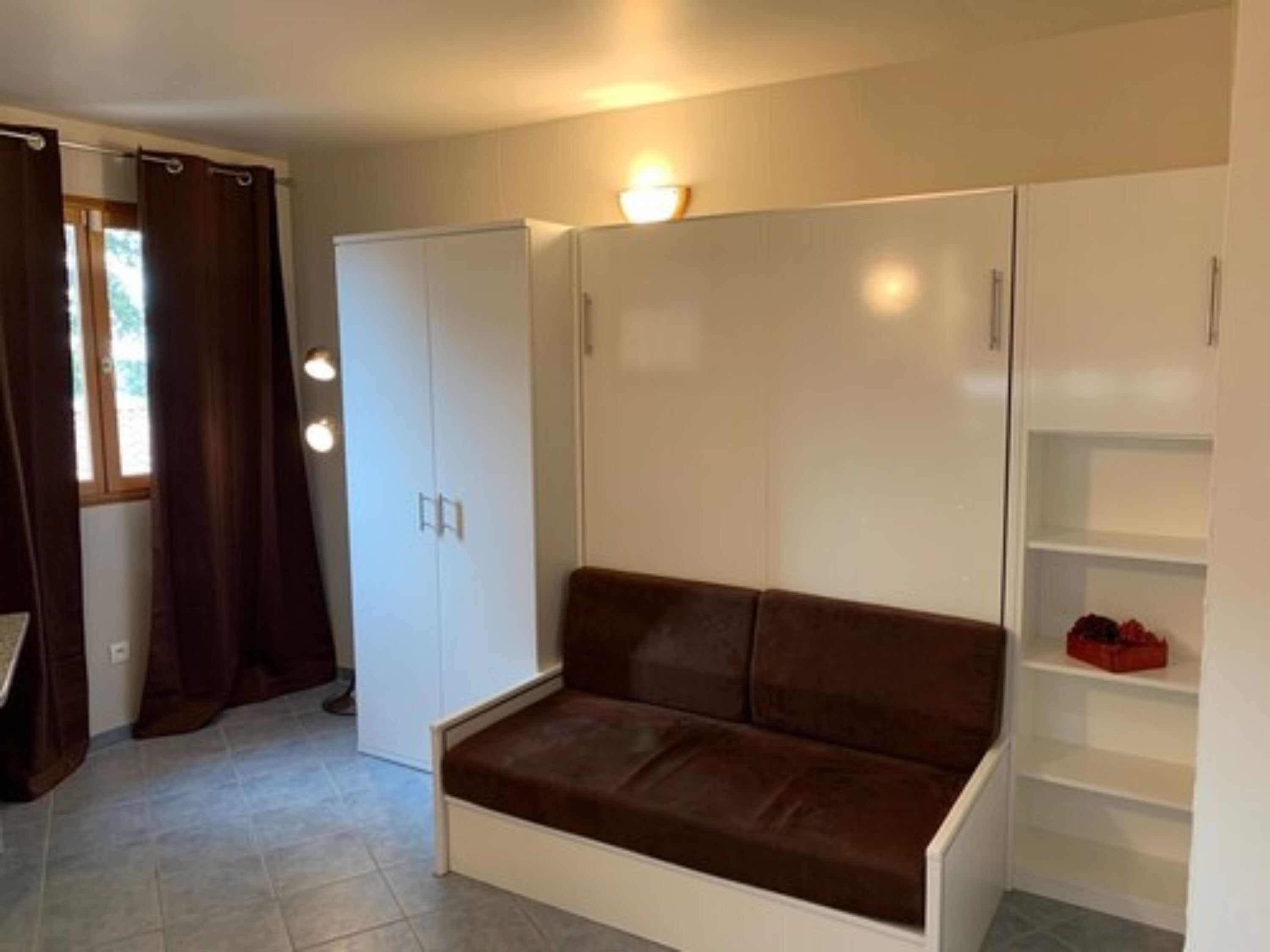 A compact room with a brown sofa, white wardrobe, and a small shelf unit.
