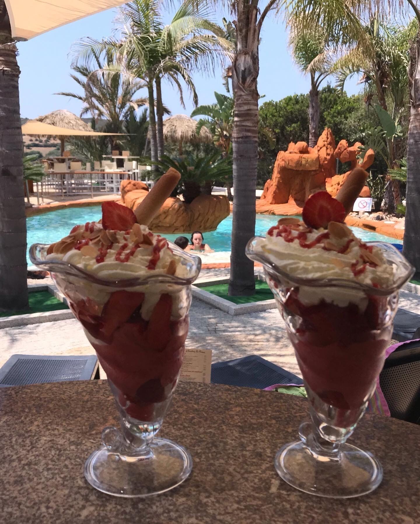 Dos vasos de postre sobre una mesa, con una piscina y palmeras al fondo.