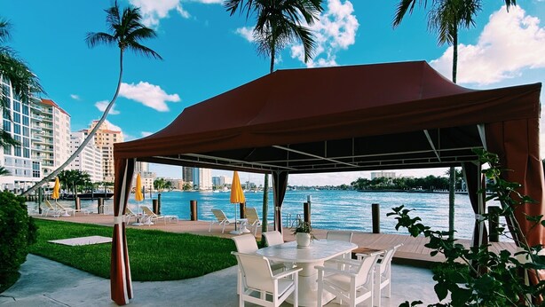 An outdoor seating area with a covered table and chairs, overlooking a body of water with palm trees and buildings in the background.