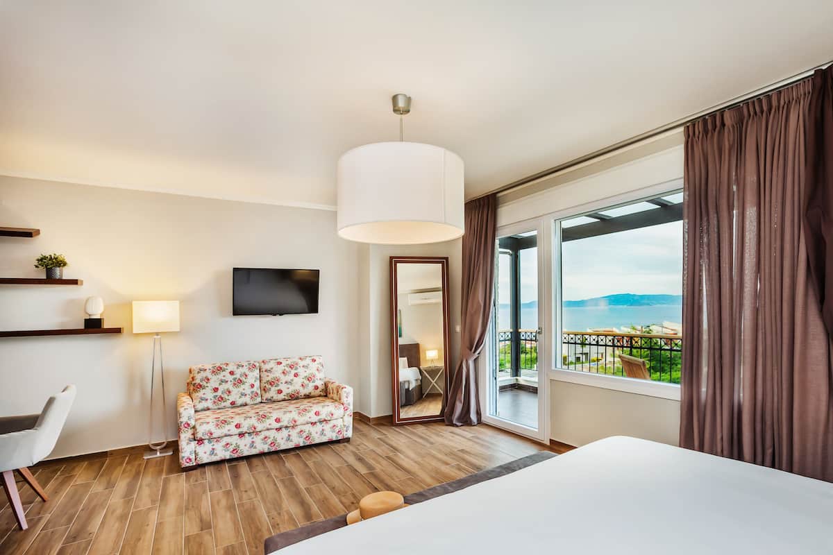 A hotel room with a wooden floor, a floral sofa, a bed, a TV, a large window with curtains, and a balcony with a view.