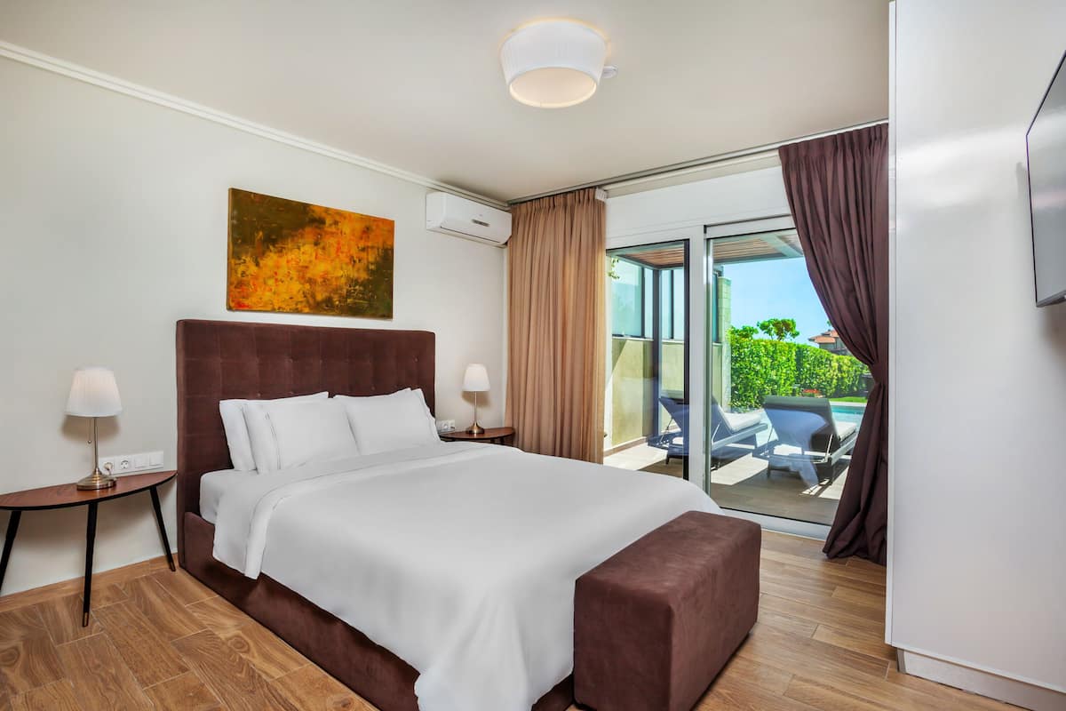 A hotel room with a large bed, a flat-screen TV, a painting on the wall, and a view of the outdoors through a sliding glass door.
