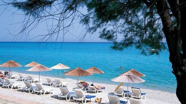 Une plage avec des chaises longues blanches et des parasols en paille, une mer d’un bleu limpide et un arbre au premier plan.