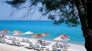 A beach with white lounge chairs and straw umbrellas, a clear blue sea, and a tree in the foreground.