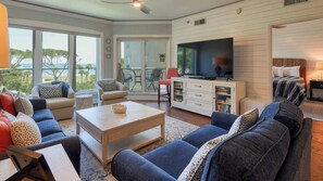 A spacious living room with a large TV, comfortable seating, a dining area, and a balcony with a view.