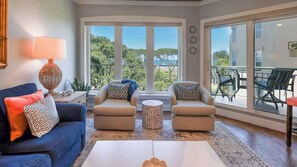 A living room with a sofa, two armchairs, a coffee table, a lamp, and a view of the outdoors through large windows.