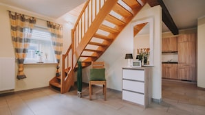 Un escalier menant à l’étage supérieur, un petit coin salon avec une chaise et une table, et un espace cuisine avec des placards en bois.