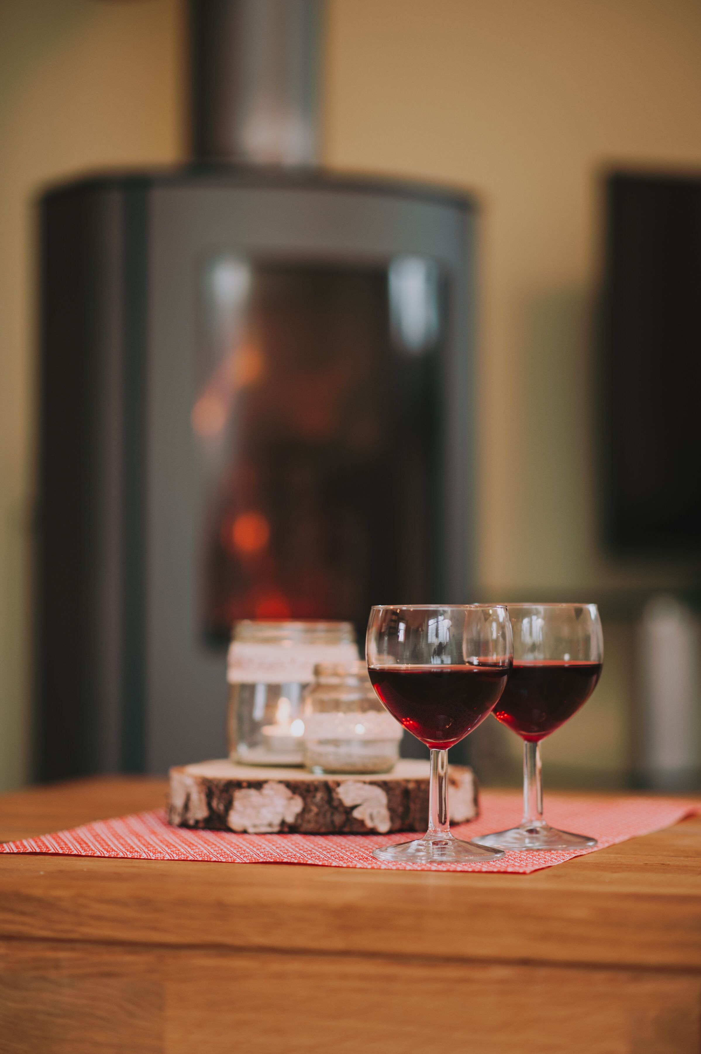 Two glasses of red wine on a wooden table with a red and white checkered placemat. In the background, there is a large, dark-colored wood-burning stove.