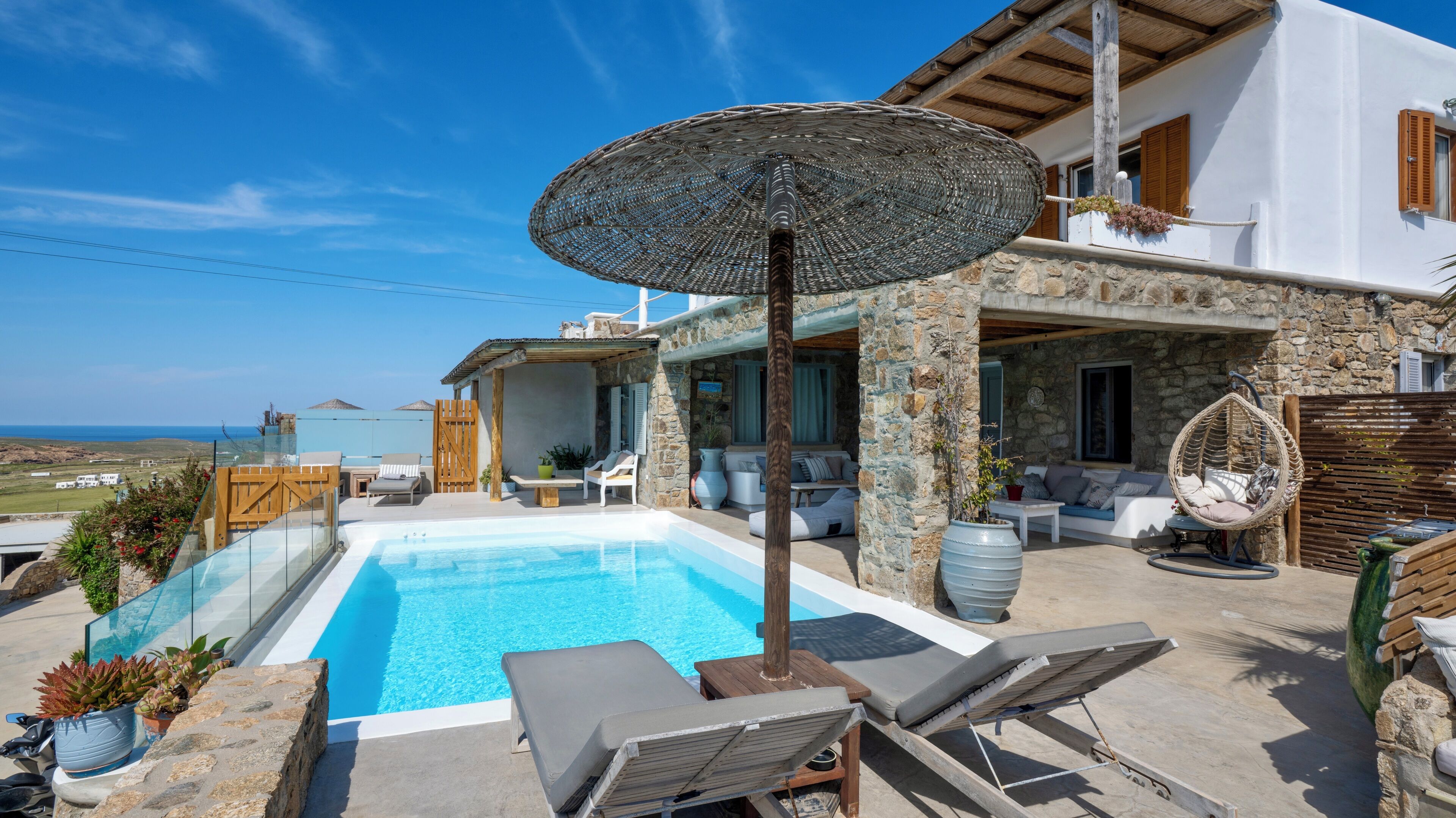 A poolside area with lounge chairs, a large umbrella, and a stone building with a view of the sea.