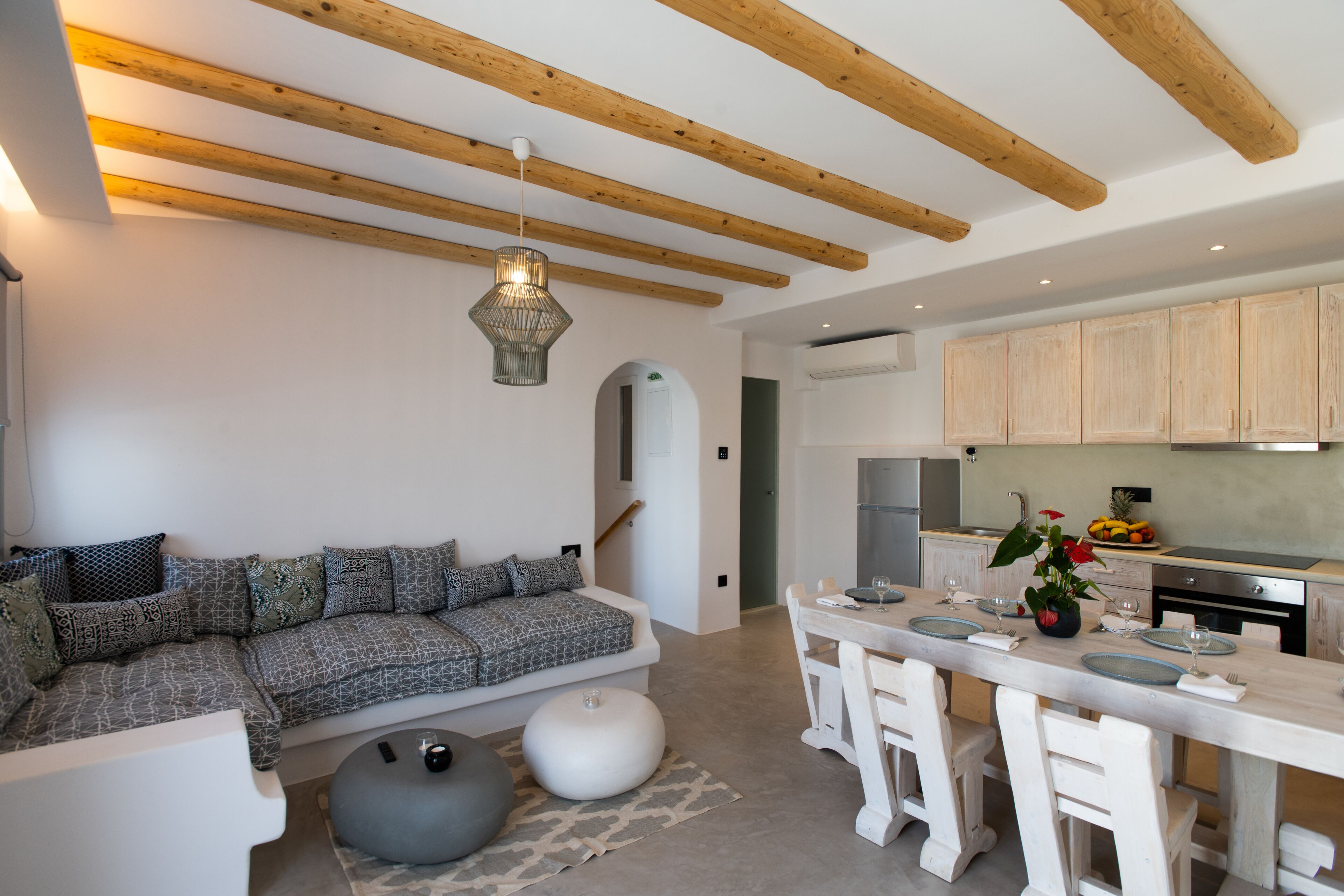 Una cocina moderna con vigas de madera, un comedor con sillas blancas y un sofá con cojines estampados.
