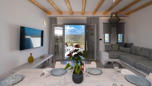 A modern dining area with a table set for four, a large flat-screen TV mounted on the wall, and a view of the outdoors through a glass door. - Mykonos Village (Mykonos)