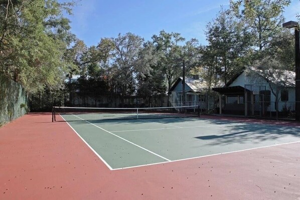 Cancha de tenis con red, rodeada de árboles y una valla.