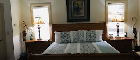 A bedroom with a wooden bed, two bedside tables with lamps, a framed artwork above the bed, and a window on each side.