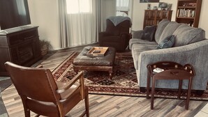 A living room with a dark brown leather sofa, a grey armchair, a wooden coffee table, and a wooden chair. There is a television, a bookshelf, and a patterned rug on the floor.