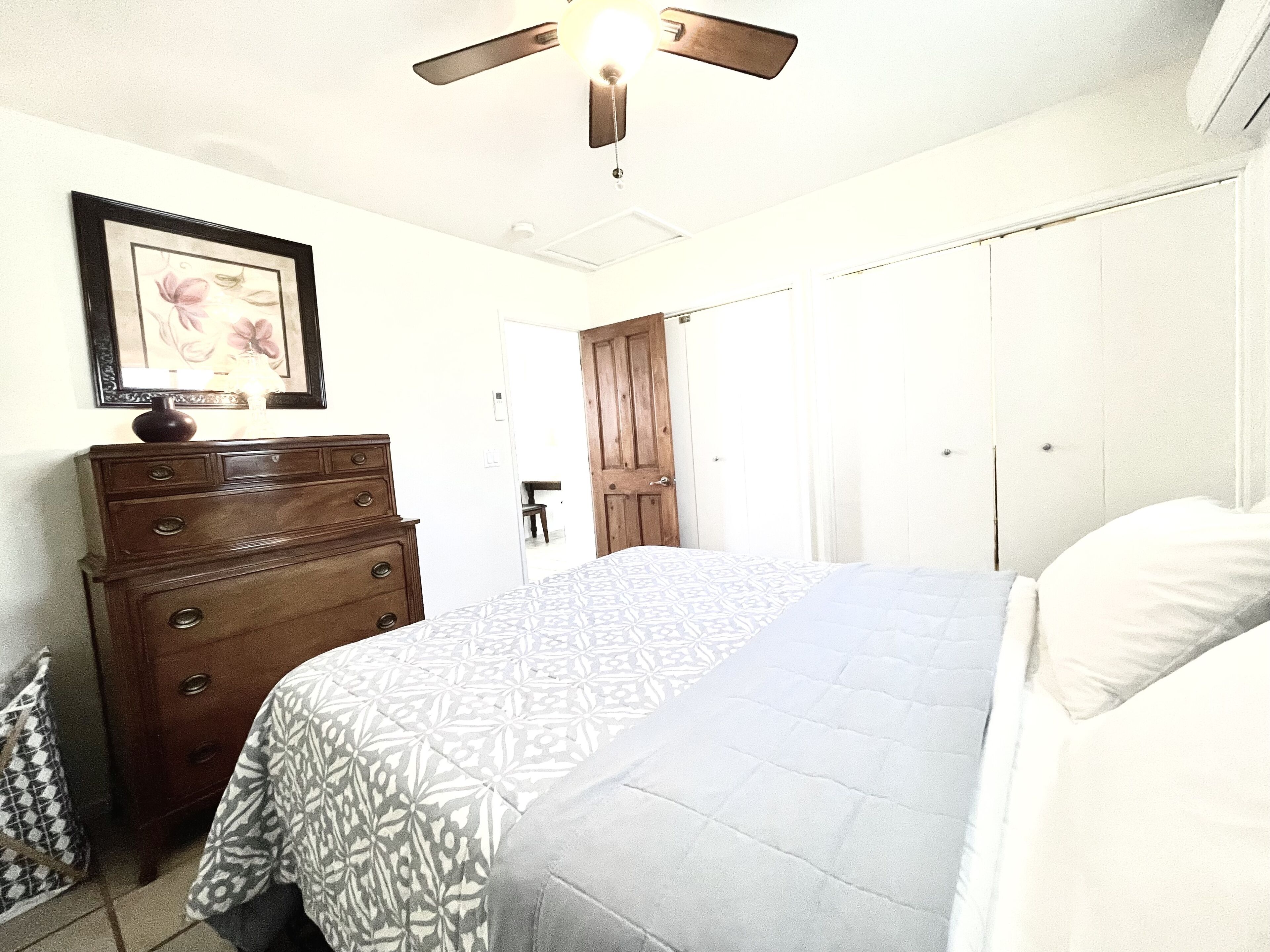 A bedroom with a bed, dresser, wardrobe, ceiling fan, and a framed picture on the wall.