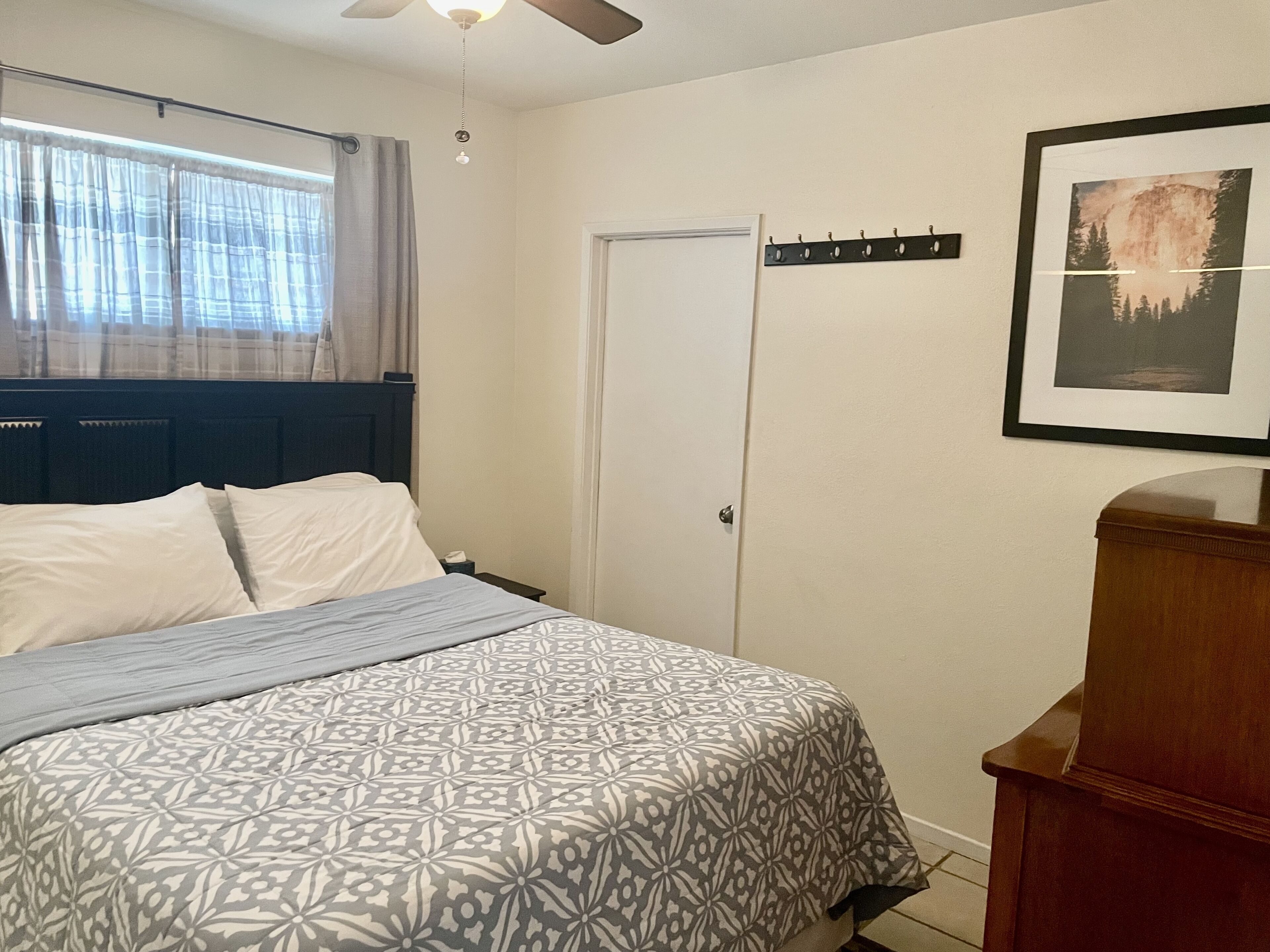 Un dormitorio con cama, ventilador de techo, puerta y perchero con un cuadro en la pared.