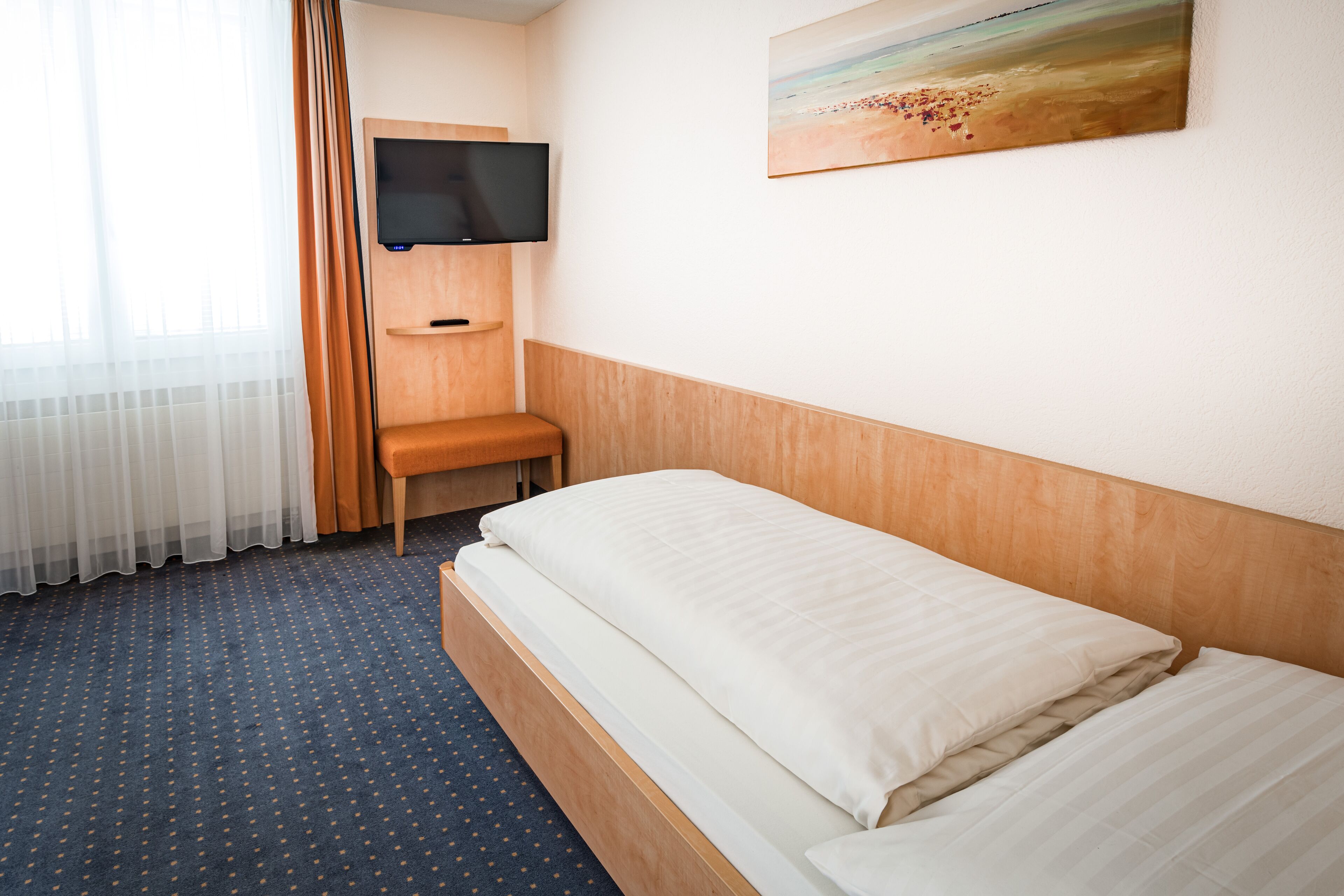 Ett hotellrum med en säng, en tv som är monterad på väggen, en tavla och ett fönster med gardiner.