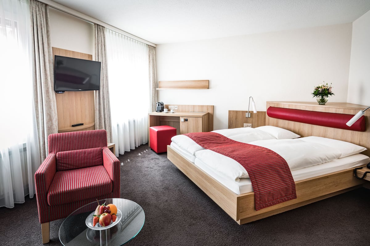 Habitación de hotel con cama, un sillón rojo, un escritorio pequeño, un televisor montado en la pared y una ventana con cortinas.