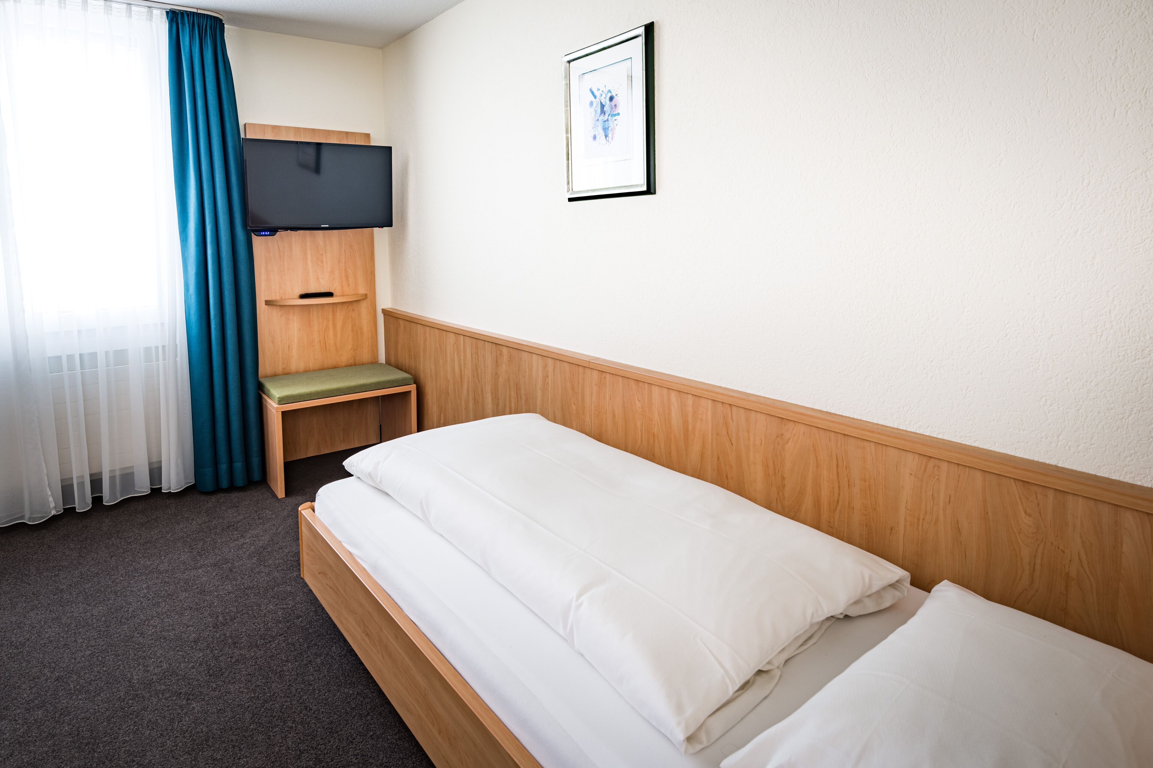 A hotel room with a single bed, a TV mounted on the wall, a small wooden shelf, and a window with curtains.
