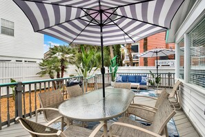 Une terrasse extérieure avec une table et des chaises en verre, un parasol à rayures et une vue sur un quartier résidentiel agrémenté de palmiers et d’une clôture blanche.