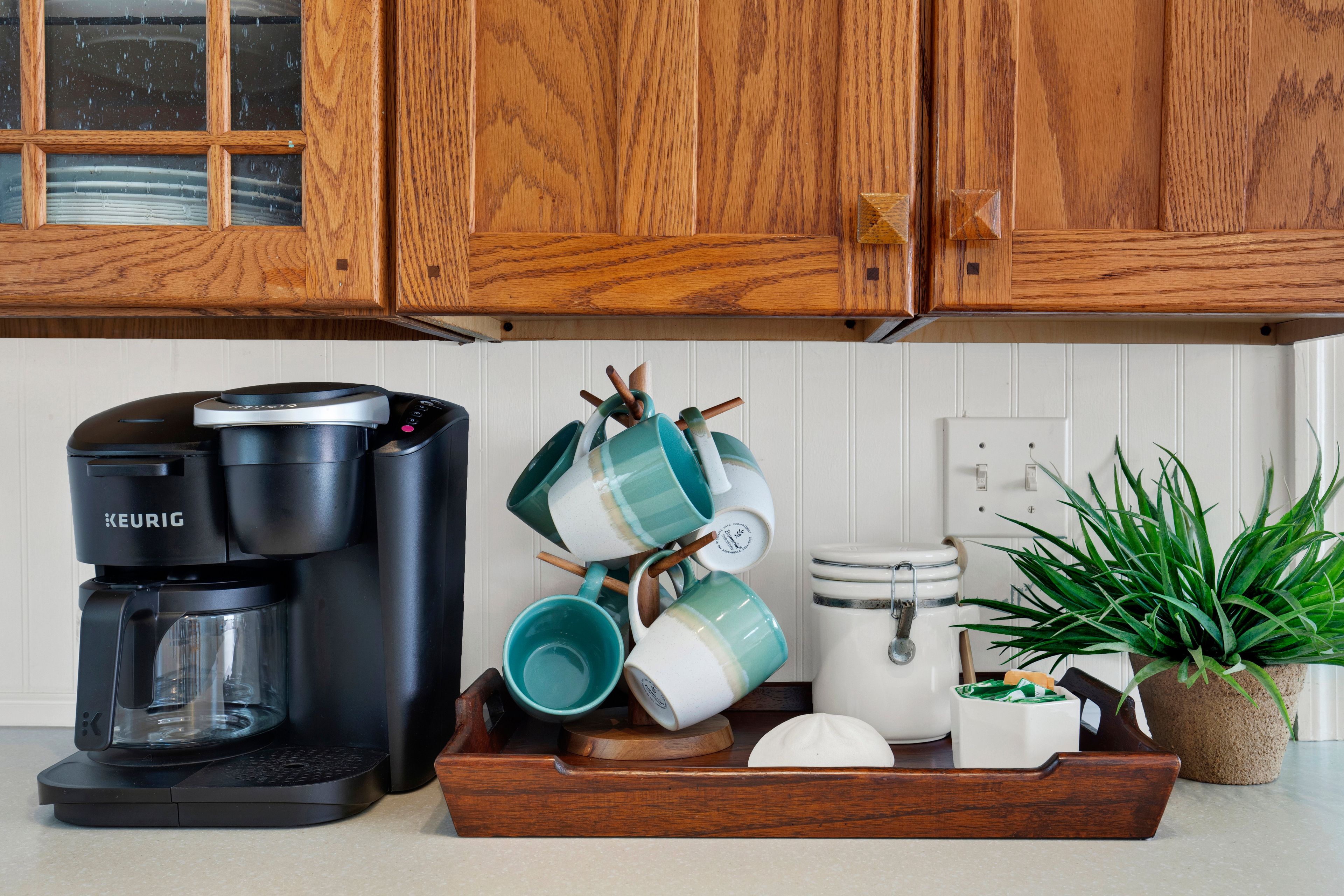 A kitchen with wooden cabinets, a Keurig coffee maker, a tray with mugs, a white appliance, and a potted plant.